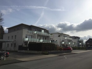 A row of modern multi-story residential buildings with balconies, a clear sky, and a red car parked on the street.