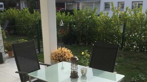 An outdoor patio with a glass table and two chairs, surrounded by a garden and residential buildings. - Boardinghouse Oelde - Apartment mit Terrasse (Oelde)