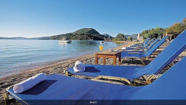 Ein Strand mit Liegestühlen, einem Tisch mit Handtuch und einem Orangensaftglas, und ein Boot im Wasser.