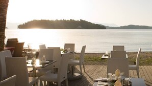 Ein Restaurant am Meer mit Außenbestuhlung und Blick auf das Wasser, ein Boot und eine Person am Ufer.