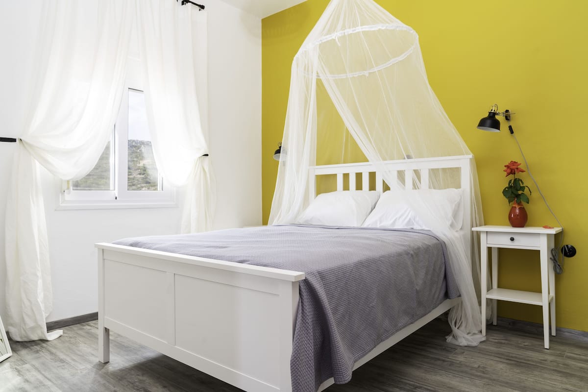 A white bed with a grey bedspread, a white nightstand with a vase and a lamp, and a yellow wall.