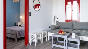 A modern living room with a grey sofa, a small white table with a fruit bowl, and a red-framed window with white curtains.
