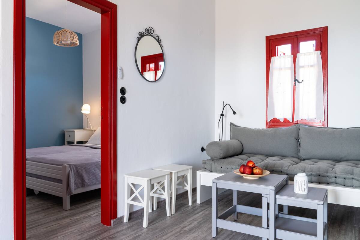 A modern living room with a grey sofa, a small white table with a fruit bowl, and a red-framed window with white curtains.