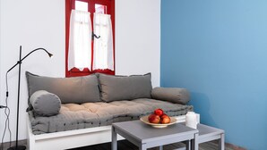 A modern living room with a grey sofa, a small table with a bowl of apples, a floor lamp, and a window with red curtains.
