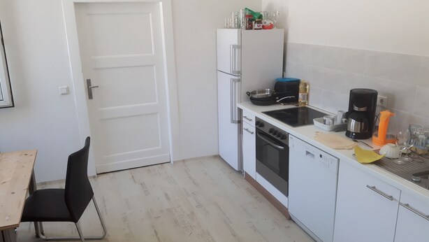 Eine kleine Küche mit einem weißen Kühlschrank, Herd und Geschirrspüler. Ein Holztisch und ein schwarzer Stuhl sind vorhanden.