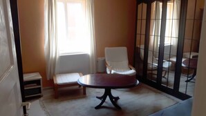 Ein Zimmer mit weißem Stuhl, kleinem Tisch, Heizkörper und einem Fenster mit Vorhängen.
