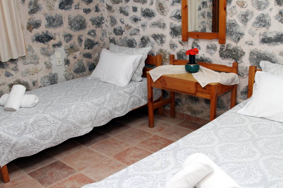 A room with two single beds, a wooden table with a vase of flowers, and a stone wall.