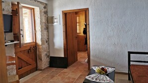 A room with a wooden door, a small table with a fruit bowl, a television on a shelf, and a ceiling light. - Cavo Ponta (Gortyna)
