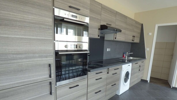 A modern kitchen with a built-in oven, microwave, and washing machine, featuring light wood cabinetry and dark grey tiles.