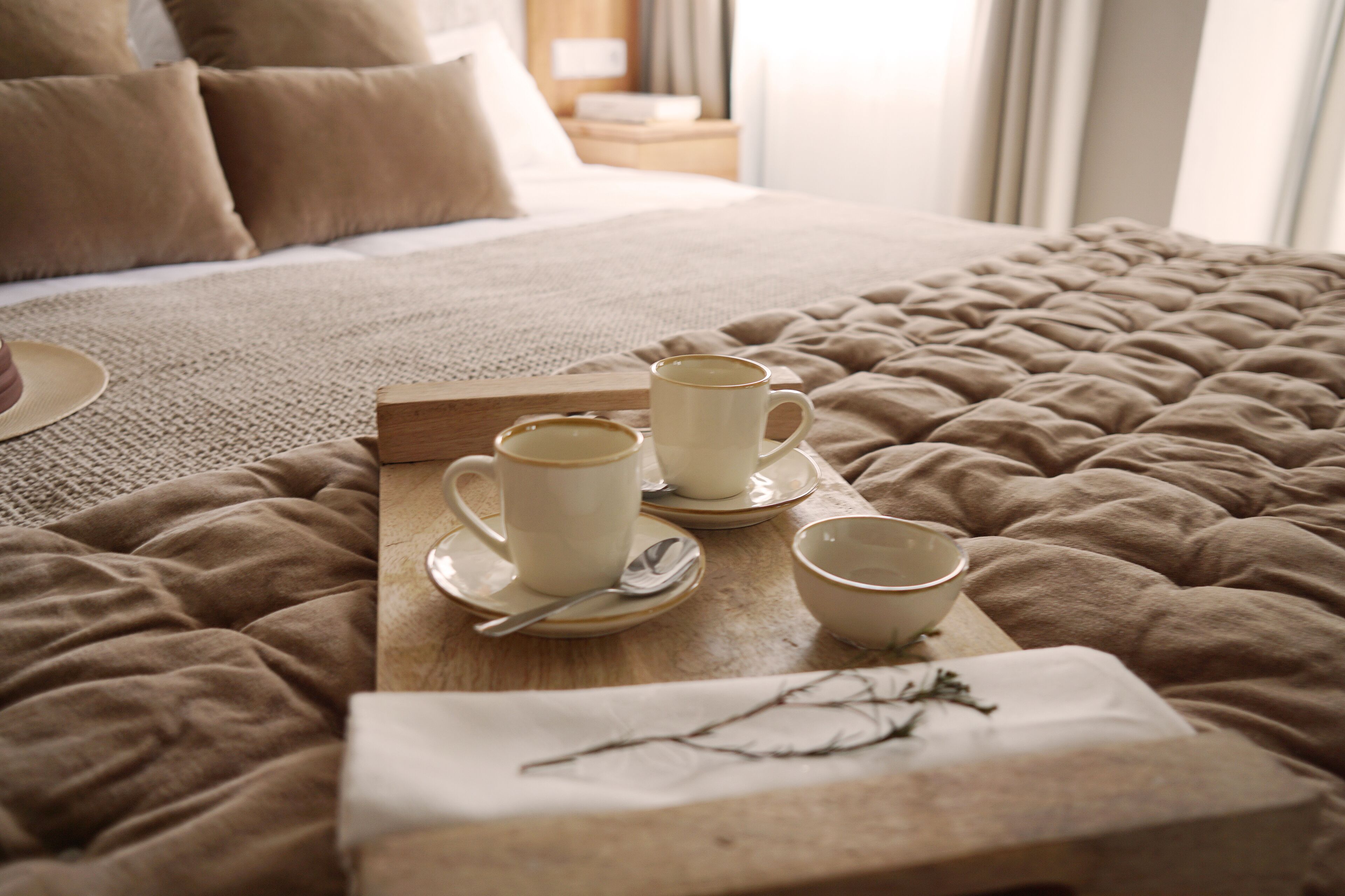 A neatly made bed with a beige quilt, a tray with three cups and a spoon, and a branch on a napkin.
