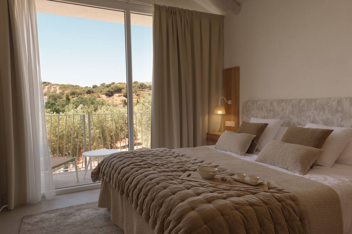 Une chambre avec un grand lit, une table de chevet avec une lampe et une vue sur une colline, visible à travers une porte-fenêtre coulissante.