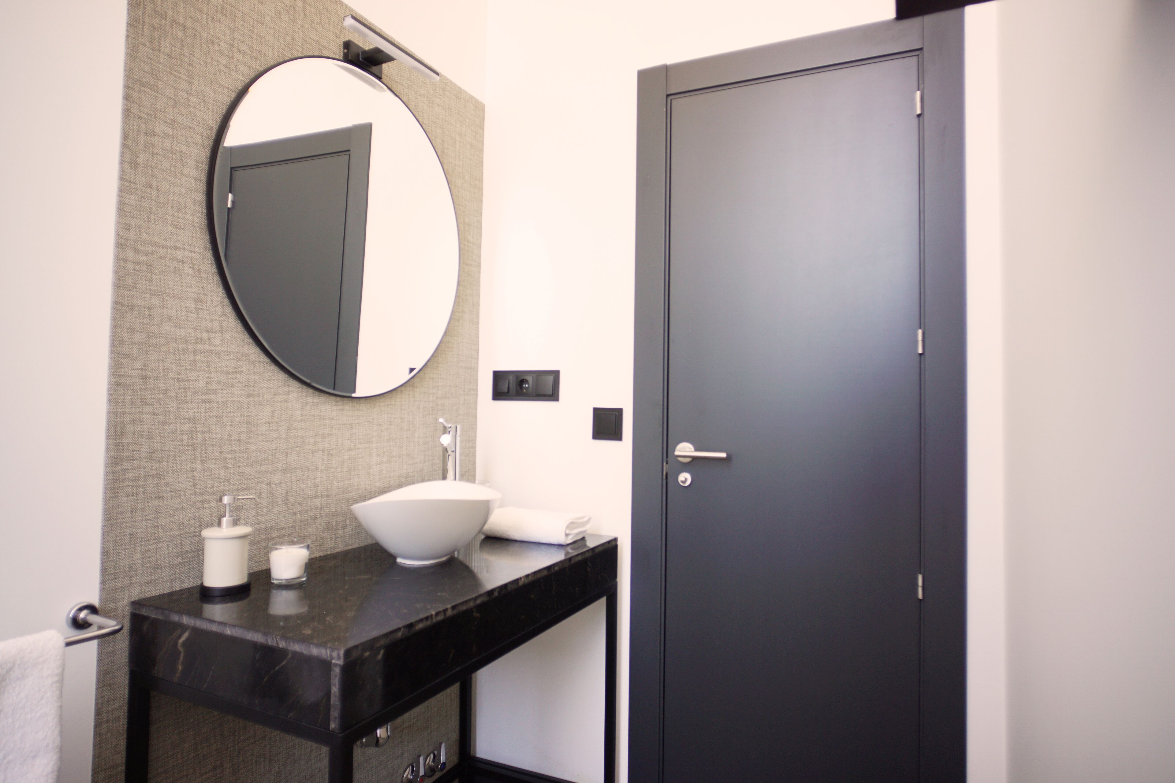 A modern bathroom with a black vanity, a round mirror, a white vessel sink, and a dark door.