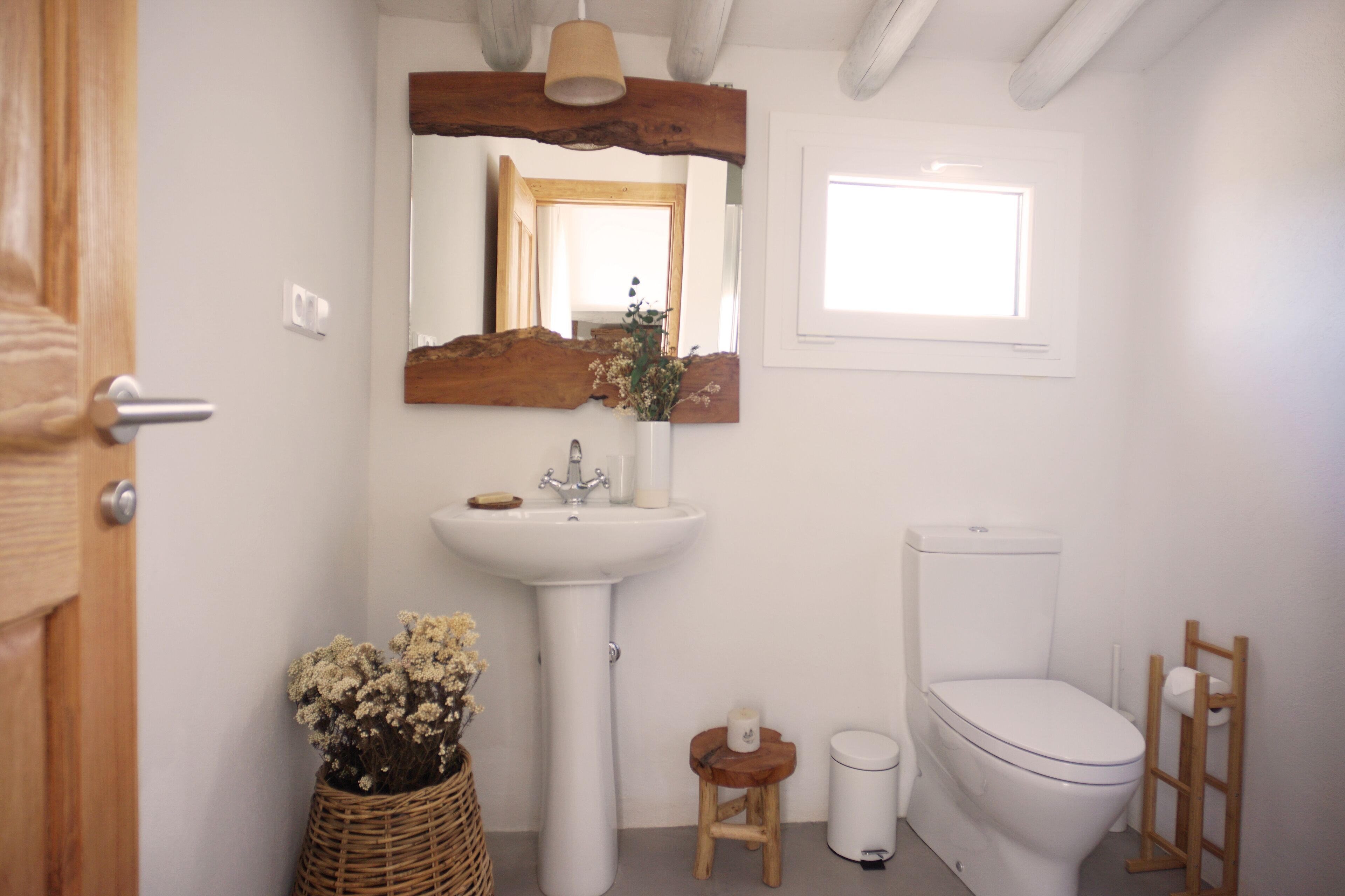 A bathroom with a white sink, mirror, and toilet. There is a wooden stool and a wicker basket with flowers.