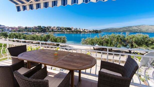 Una terraza con mesa de madera y sillas de mimbre, con vistas a la costa, edificios y árboles.