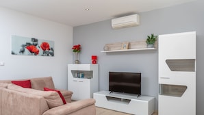 Un salón moderno con un sofá color beige, un televisor de pantalla plana, muebles blancos y un jarrón rojo con flores.