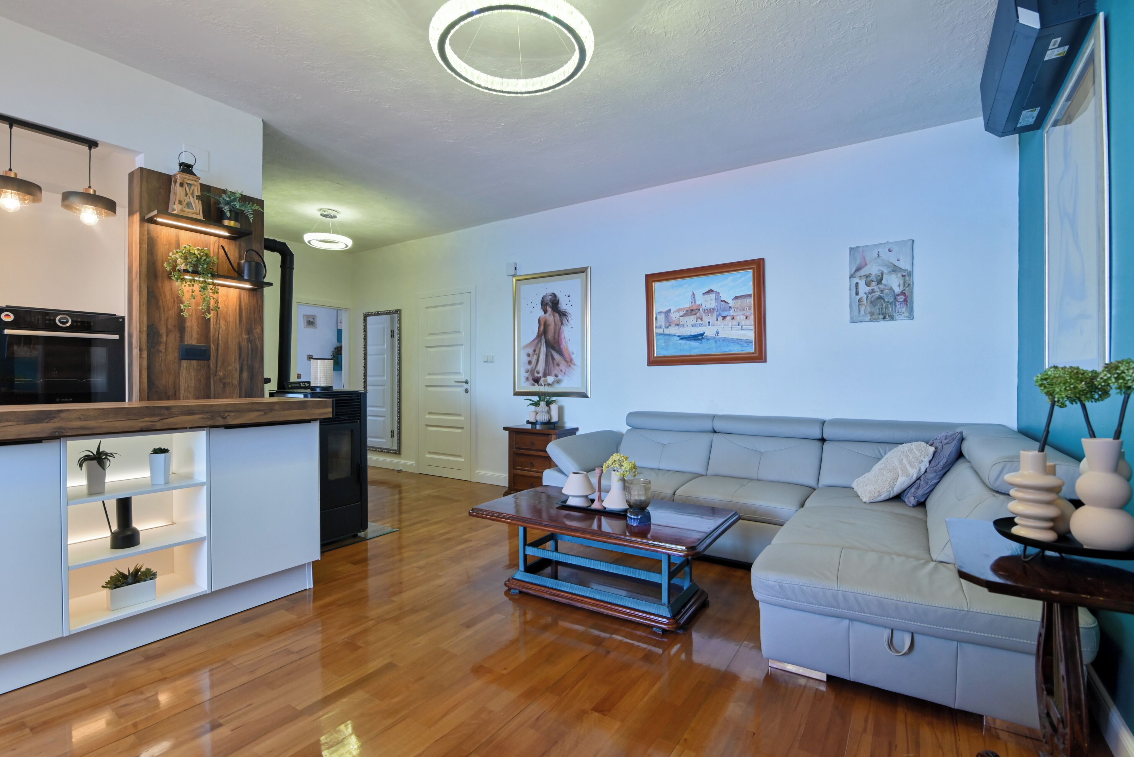 A modern living room with a sofa, coffee table, and a kitchen area with a built-in oven and shelves.