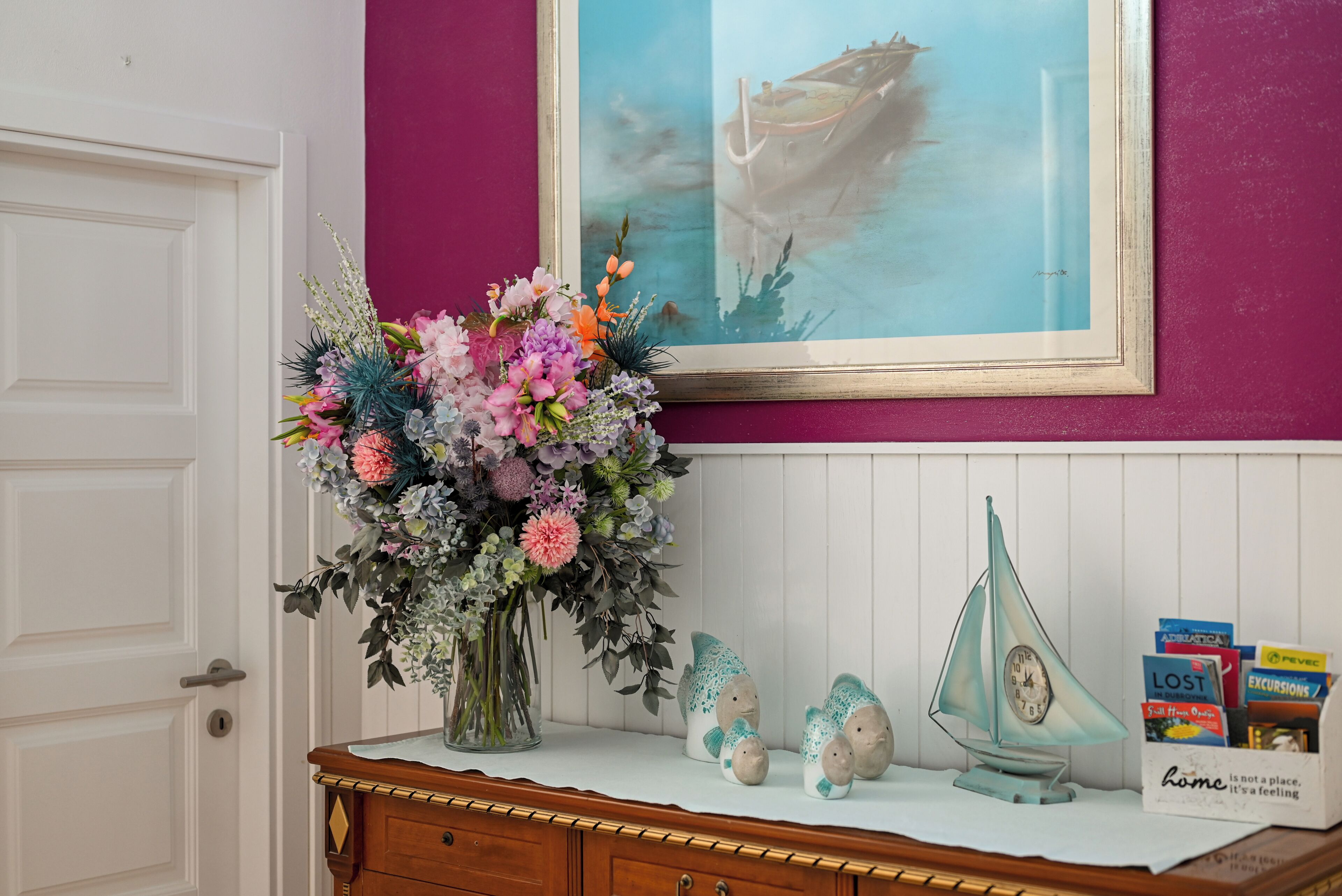 A room with a white door, a purple wall, a framed painting of a boat, a vase of flowers, decorative fish figurines, and a small sailboat model.