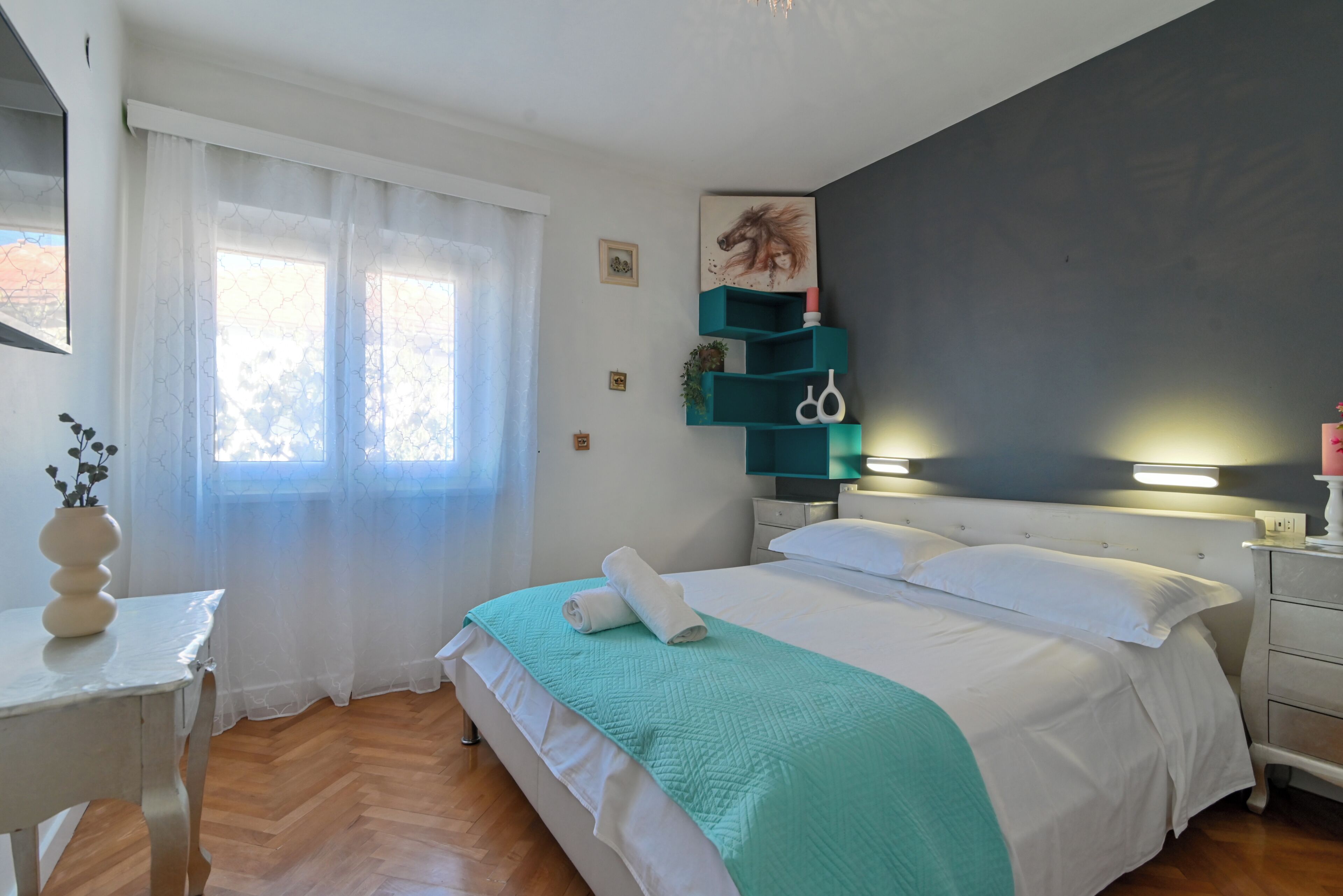 Un dormitorio ordenado con cama, mesita de noche, ventana con cortinas y estante en la pared con objetos decorativos.