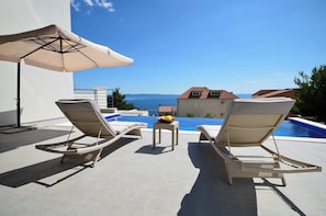 A poolside area with lounge chairs and an umbrella, overlooking a swimming pool and a view of the sea. - Villa Nina Makarska With Heated Infinity Pool of 33 m2 and Stunning sea Views (Makarska)