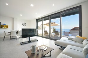 Un salon moderne doté d’un téléviseur à écran plat, d’un coin repas avec des chaises, d’une table basse avec une bouilloire et des tasses, et d’une grande porte-fenêtre coulissante donnant sur un balcon aménagé avec des sièges d’extérieur.