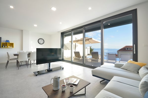 A modern living room with a flat-screen TV, a dining area with chairs, a coffee table with a kettle and cups, and a large sliding glass door leading to a balcony with outdoor seating. - Villa Nina Makarska With Heated Infinity Pool of 33 m2 and Stunning sea Views (Makarska)