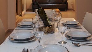 A dining table set for six with plates, cutlery, and wine glasses. A bottle of wine is placed in the center. The room has a sofa and a colorful painting on the wall. - Citybreak Apartman With Private Beach Near Split (Omis)