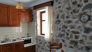 A kitchen with wooden cabinets, a white tiled backsplash, a stone wall, a window with curtains, a table with a tablecloth, and a clock on the wall. - Nefeli's traditional suite no.12 (Gortyna)