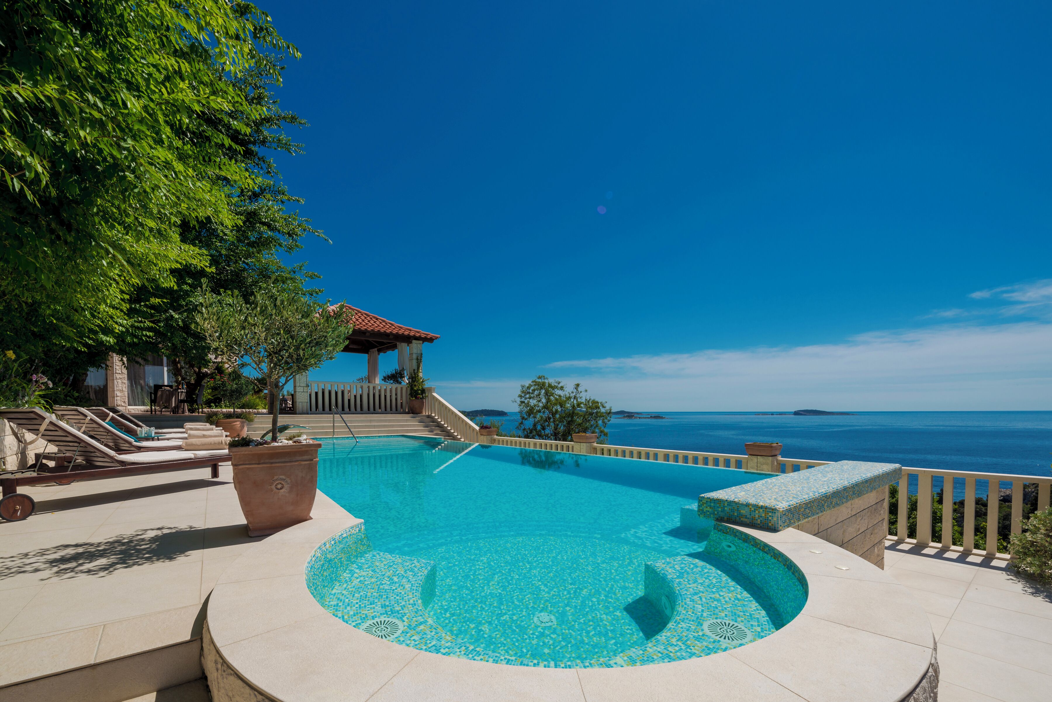 A pool with a tiled edge, a lounge area with chairs, and a view of the sea.