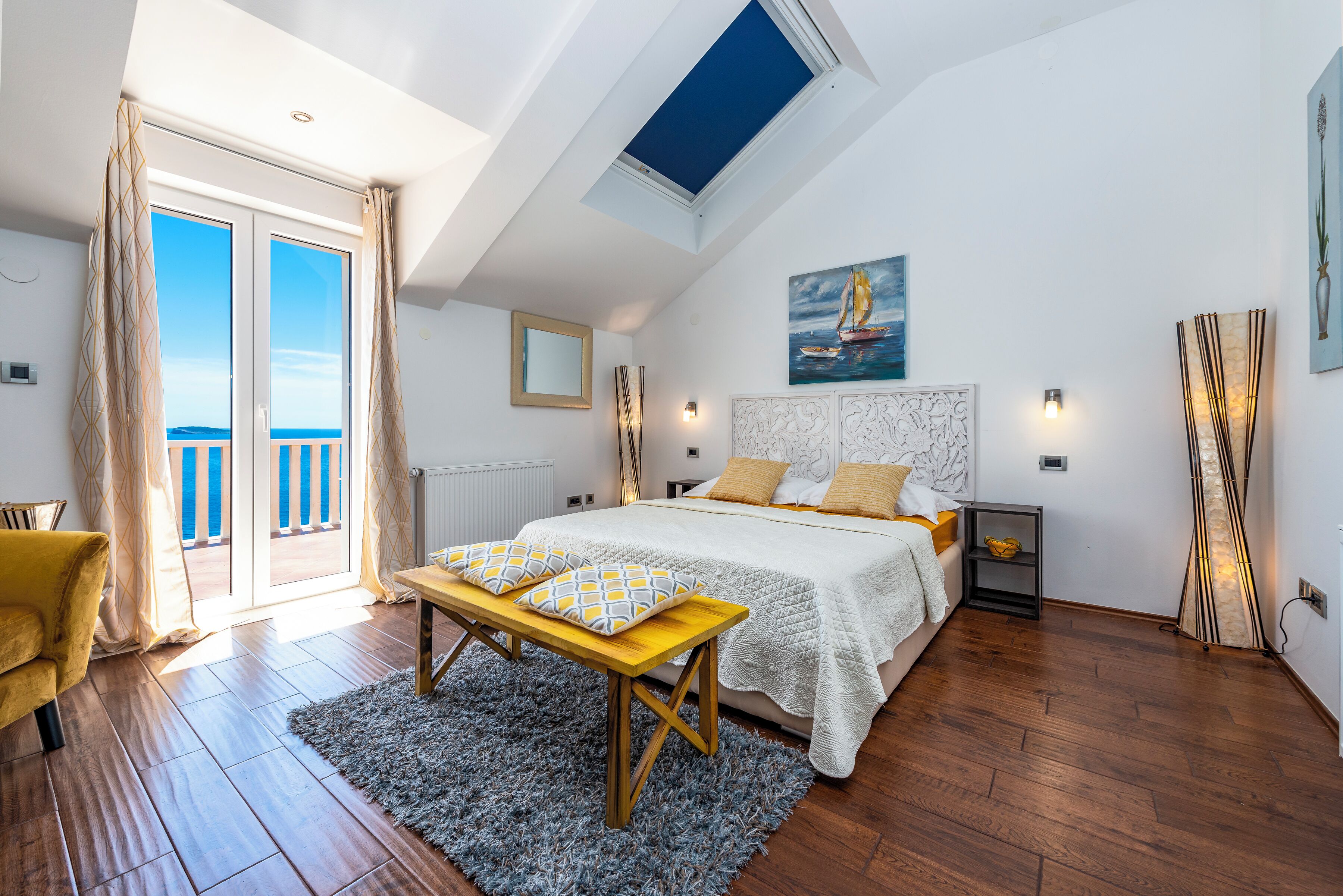 Ein Schlafzimmer mit Dachfenster, einem Bett mit weißer Daunendecke, einer Holzbank und Meerblick vom Balkon.