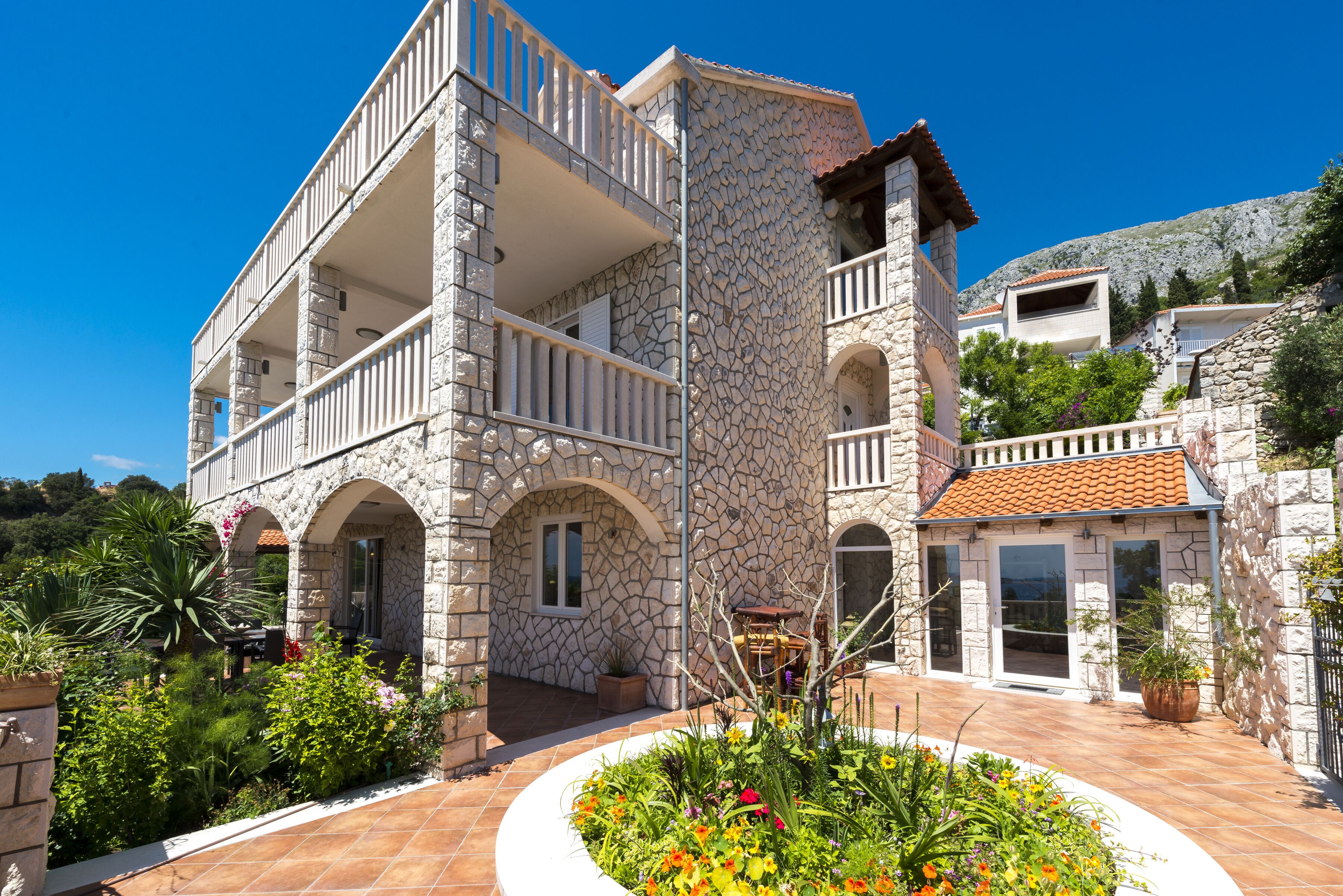 A two-story stone building with balconies, a garden with various plants, and a clear blue sky.
