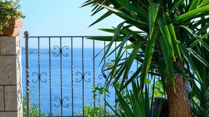 Een balkon met een sierhek van smeedijzer, uitzicht op zee en weelderige begroeiing.