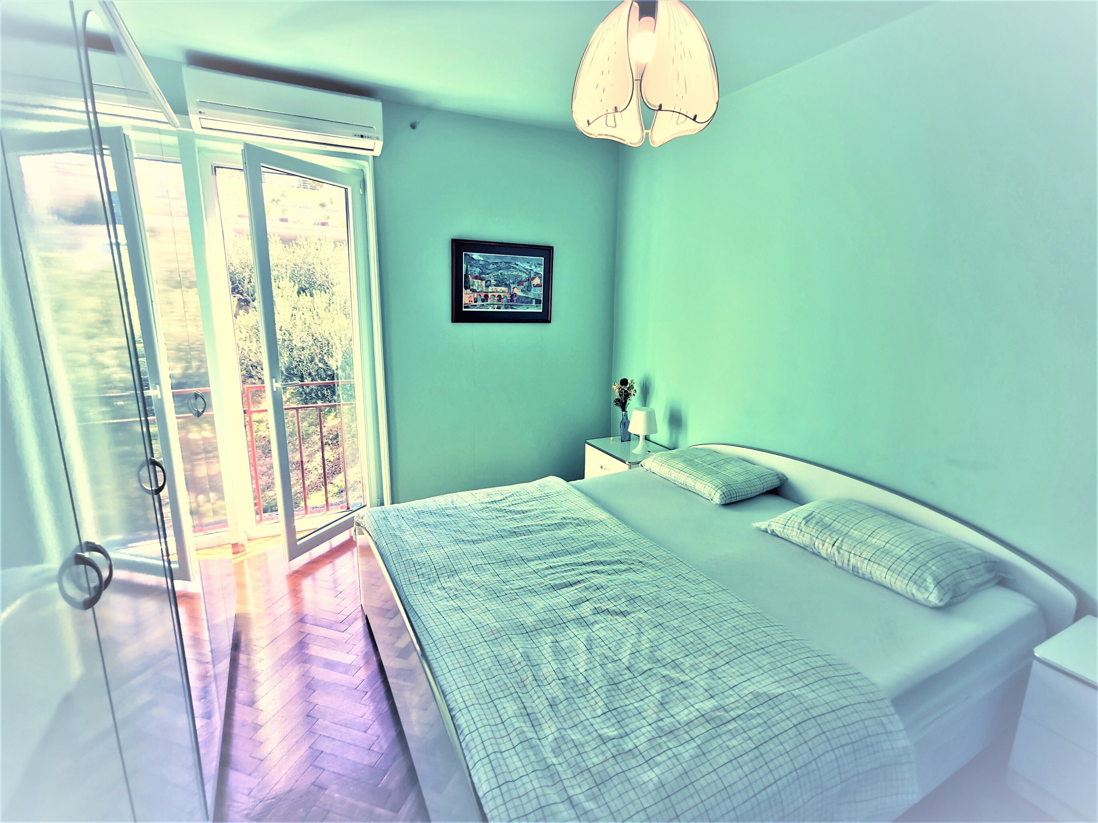 A bedroom with a bed, a nightstand, a lamp, a framed picture, and a view through a glass door.