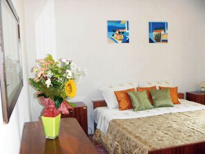 A bedroom with a bed, pillows, a nightstand, a bedside table, a vase with flowers, and two wall art pieces.
