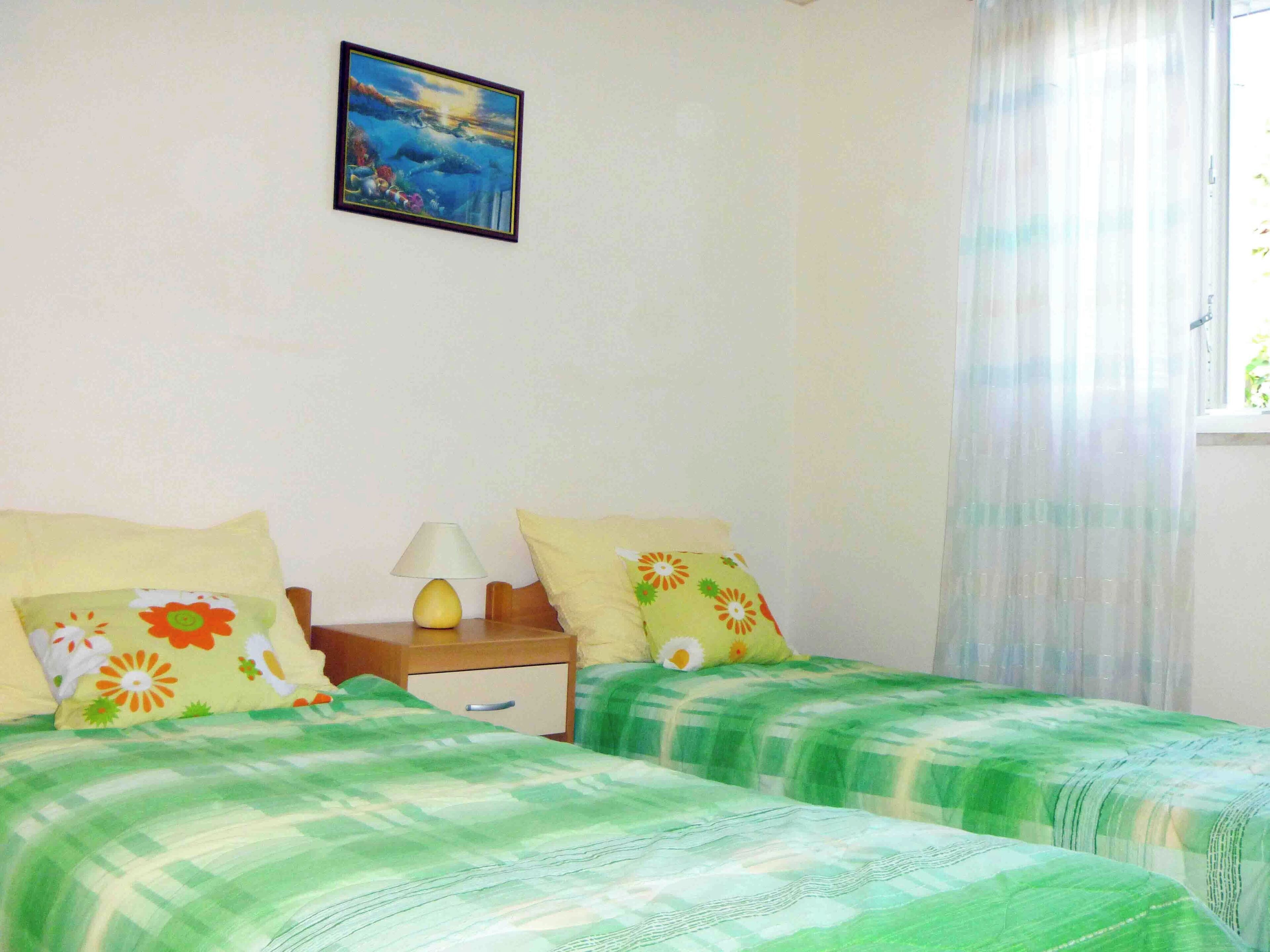A bedroom with two beds, a nightstand, a lamp, a window with sheer curtains, and a painting on the wall.