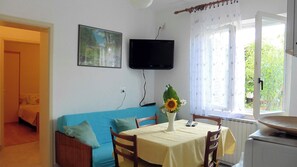 A room with a blue sofa, a table with a yellow tablecloth, a TV mounted on the wall, and a window with curtains.