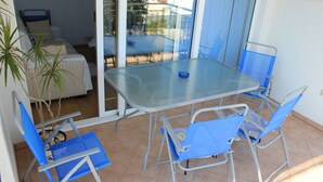 Ein Glastisch mit vier blauen Stühlen, ein kleines blaues Objekt auf dem Tisch und eine Schiebetür zu einer Terrasse mit einem Bett und einer Pflanze.