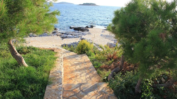 Ein steiniger Weg führt zu einer felsigen Küstenlinie mit Blick auf das Meer und eine entfernte Insel.
