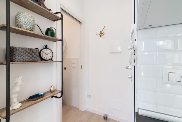 A bathroom with a white tiled wall, a white toilet, a white sink, and a white shower. There is a shelf with a clock, a small statue, and a green watering can.
