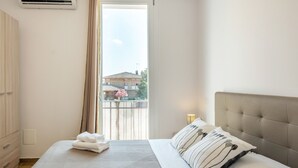 Een slaapkamer met een bed, een kledingkast en een raam met uitzicht op gebouwen en een boom.