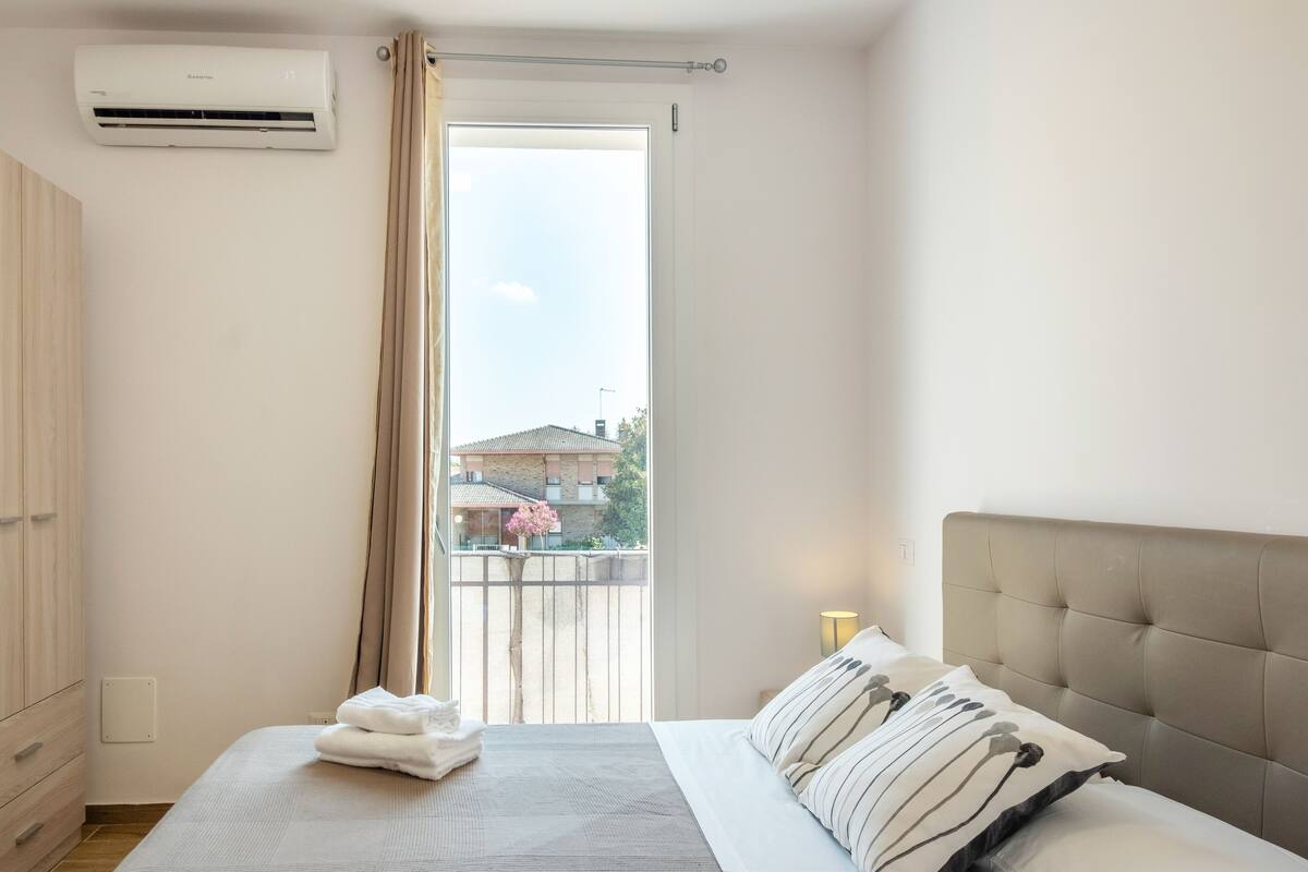 Een slaapkamer met een bed, een kledingkast en een raam met uitzicht op gebouwen en een boom.