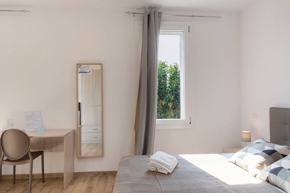Een hotelkamer met een bed, een bureau, een stoel, een raam met gordijnen en uitzicht op groen.