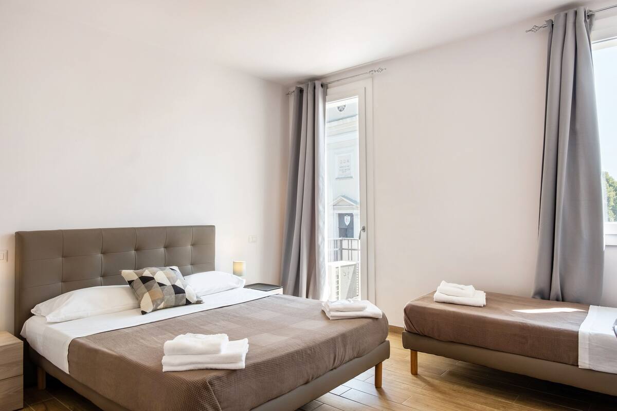 Een hotelkamer met een groot bed, een kleiner bed, een nachtkastje en een raam met gordijnen.