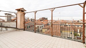 Une terrasse sur le toit, dotée d’une rambarde en métal, d’un sol carrelé et offrant une vue sur un paysage urbain avec des bâtiments et une tour.
