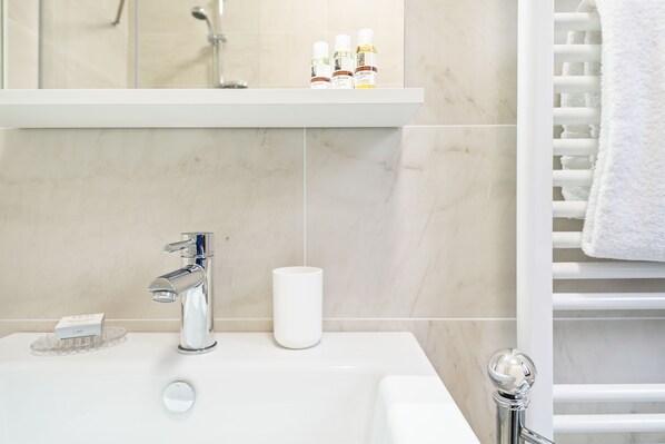 Baño con lavamanos blanco, grifo cromado y un espejo encima. Hay artículos de aseo en el lavamanos y un perchero a la derecha.