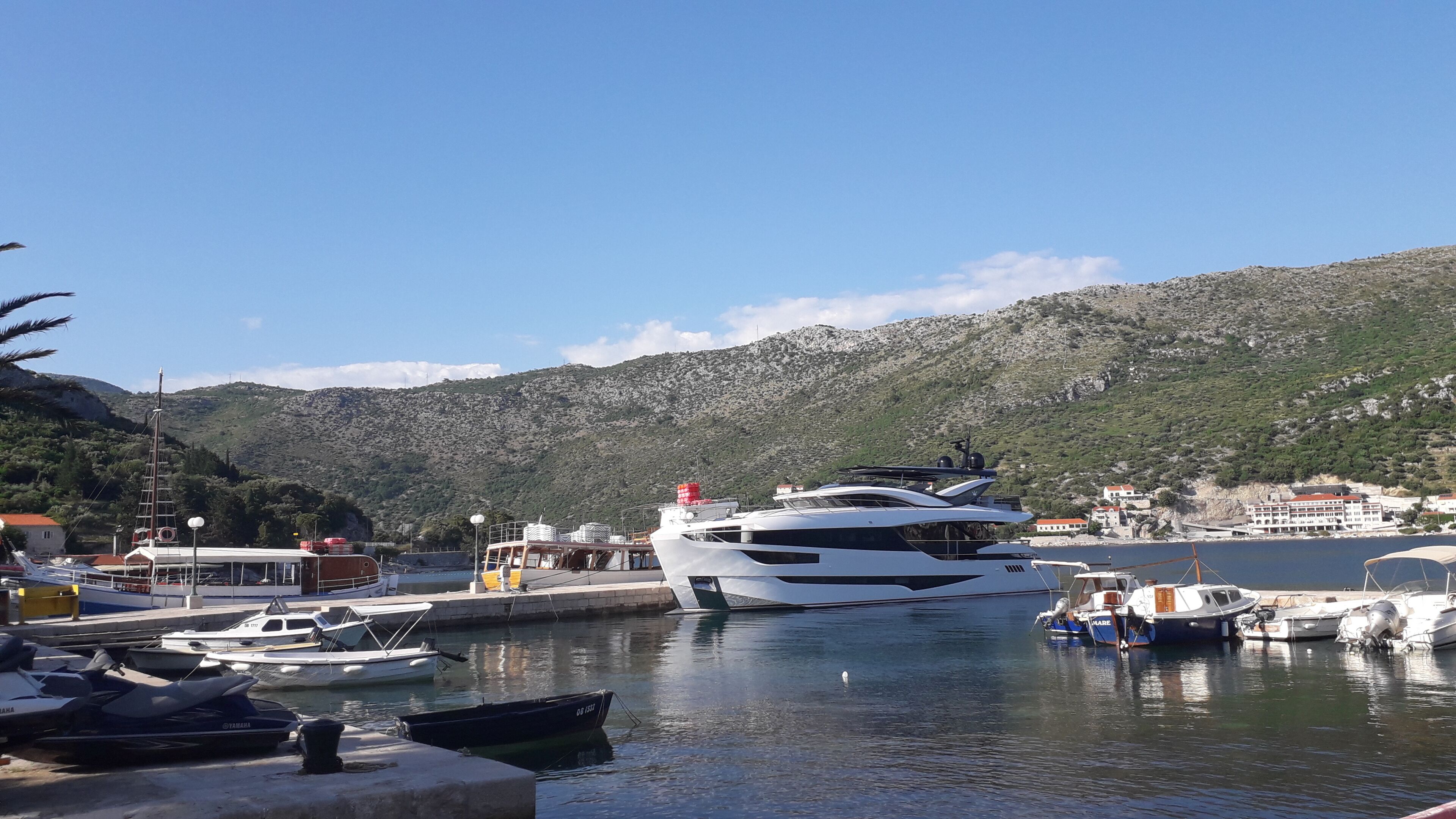 Un port de plaisance où sont amarrés différents types de bateaux, dont un grand yacht, avec en arrière-plan un paysage montagneux.
