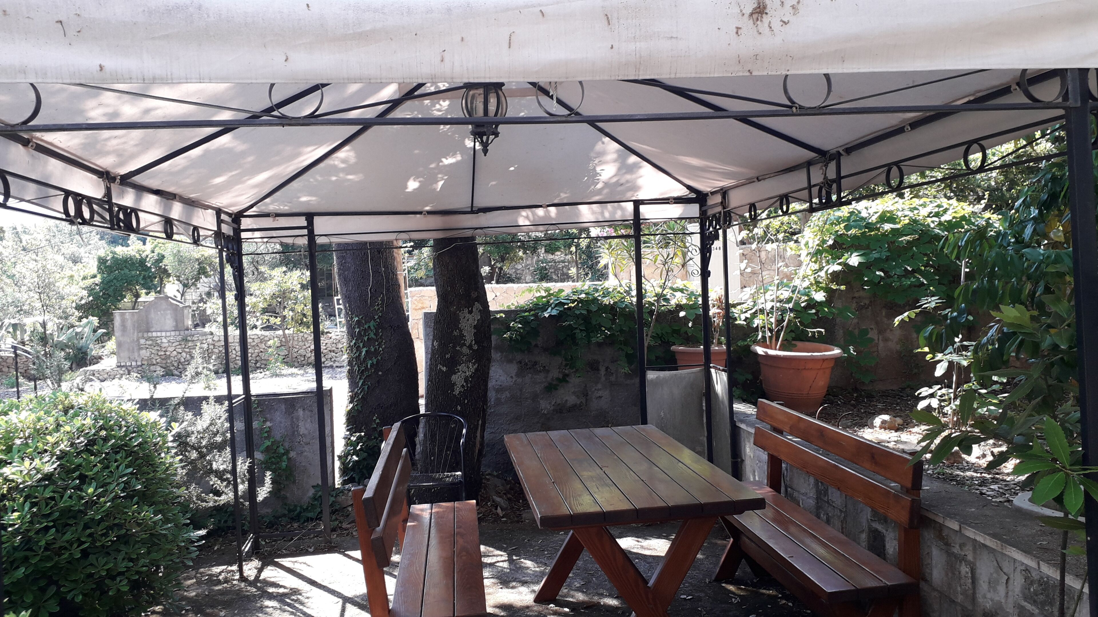 Uma área de estar coberta ao ar livre, com mesa e bancos, cercada por vegetação e um muro de pedra.