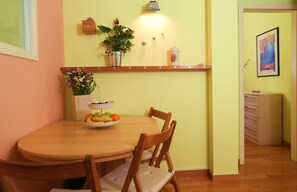 Un comedor con una mesa de madera, sillas, un frutero, una planta en maceta y un estante de pared con una lámpara.