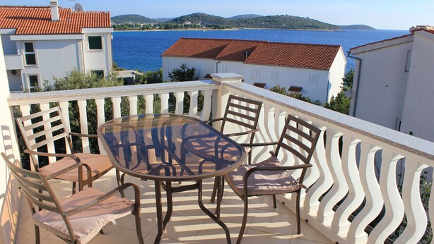 Un balcon avec une table en verre et quatre chaises, donnant sur une étendue d’eau et des collines.