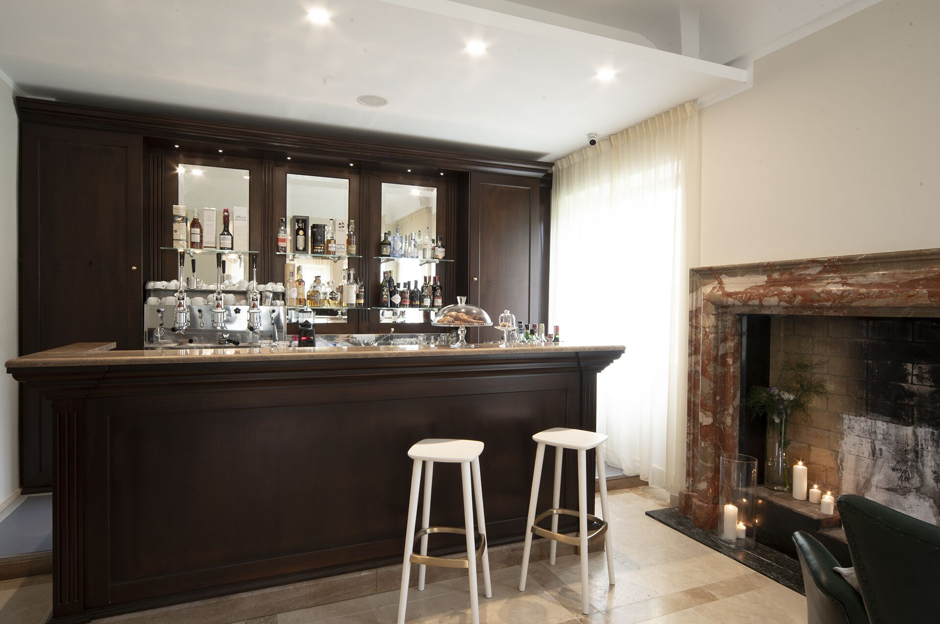 A bar area with a dark wood counter, multiple bottles, and glasses. There are two white stools in front. A fireplace with candles is visible to the right.