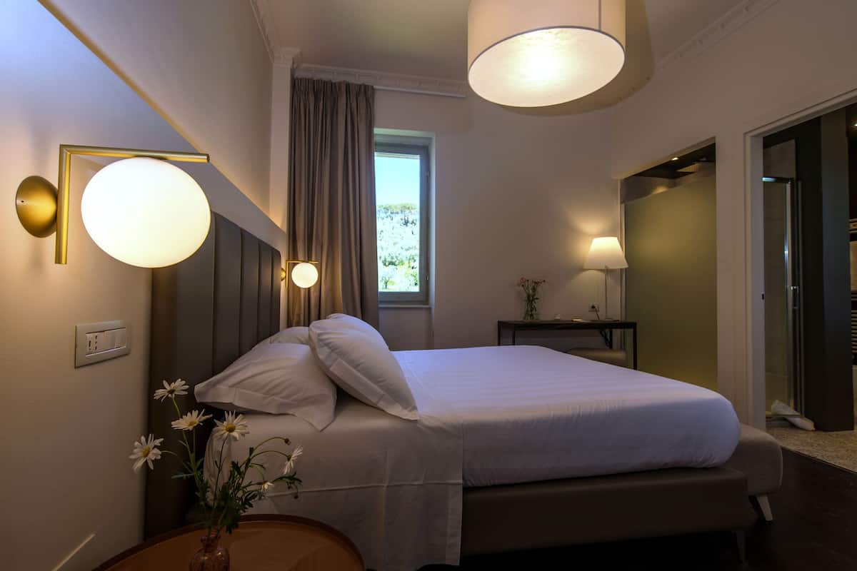 Habitación de hotel con una cama grande, lámparas de noche, una ventana con cortinas, una pequeña mesa con un florero y una puerta que da a otra habitación.
